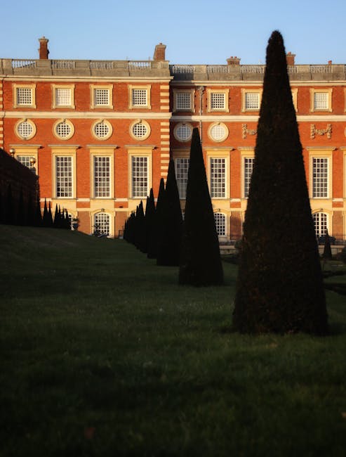 A neatly maintained formal garden area with a lush, well-trimmed green lawn and a series of tall, cone-shaped evergreen trees arranged in rows, leading towards an elegant historic building with a red-